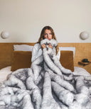 Plaid fausse fourrure sensation gris tenu dans les mains d’une femme posé dans un lit