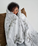 Plaid polaire gris à flocons de neige utilisé en intérieur, enveloppant une personne assise dans un fauteuil pour une ambiance cocooning et chaleureuse