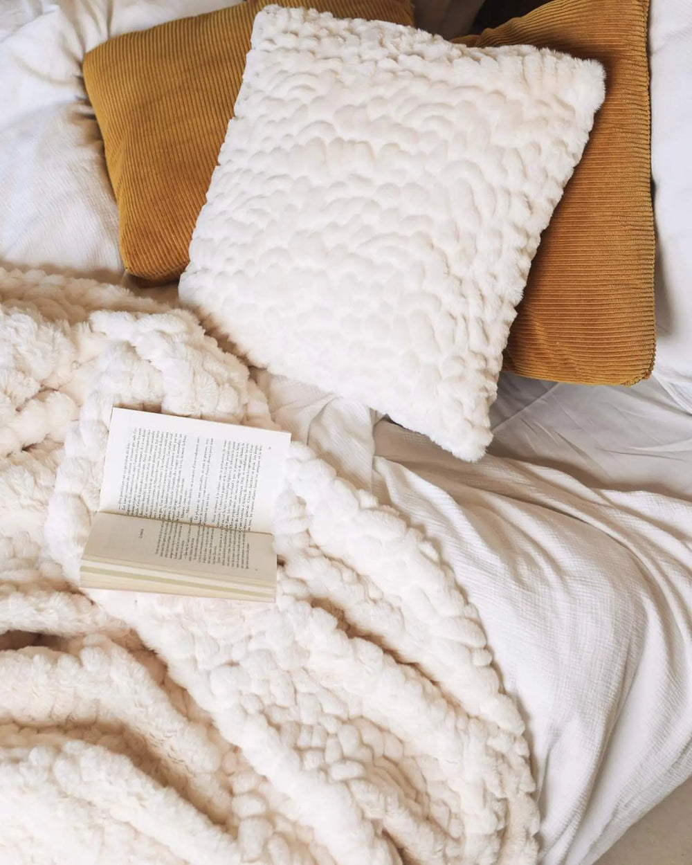 Coussin waro  carré en fausse fourrure galet blanc sur un lit 