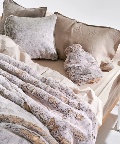 Coussin fausse fourrure Frisson Naturel waro posé sur un lit accompagné d'une bouillotte frisson et un plaid en fausse fourrure 